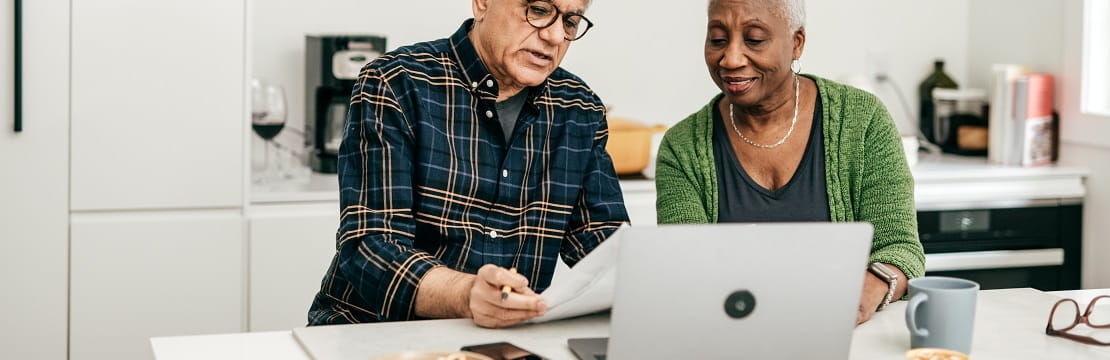 Retired couple sits at a kitchen counter reviewing documents in front of a laptop.